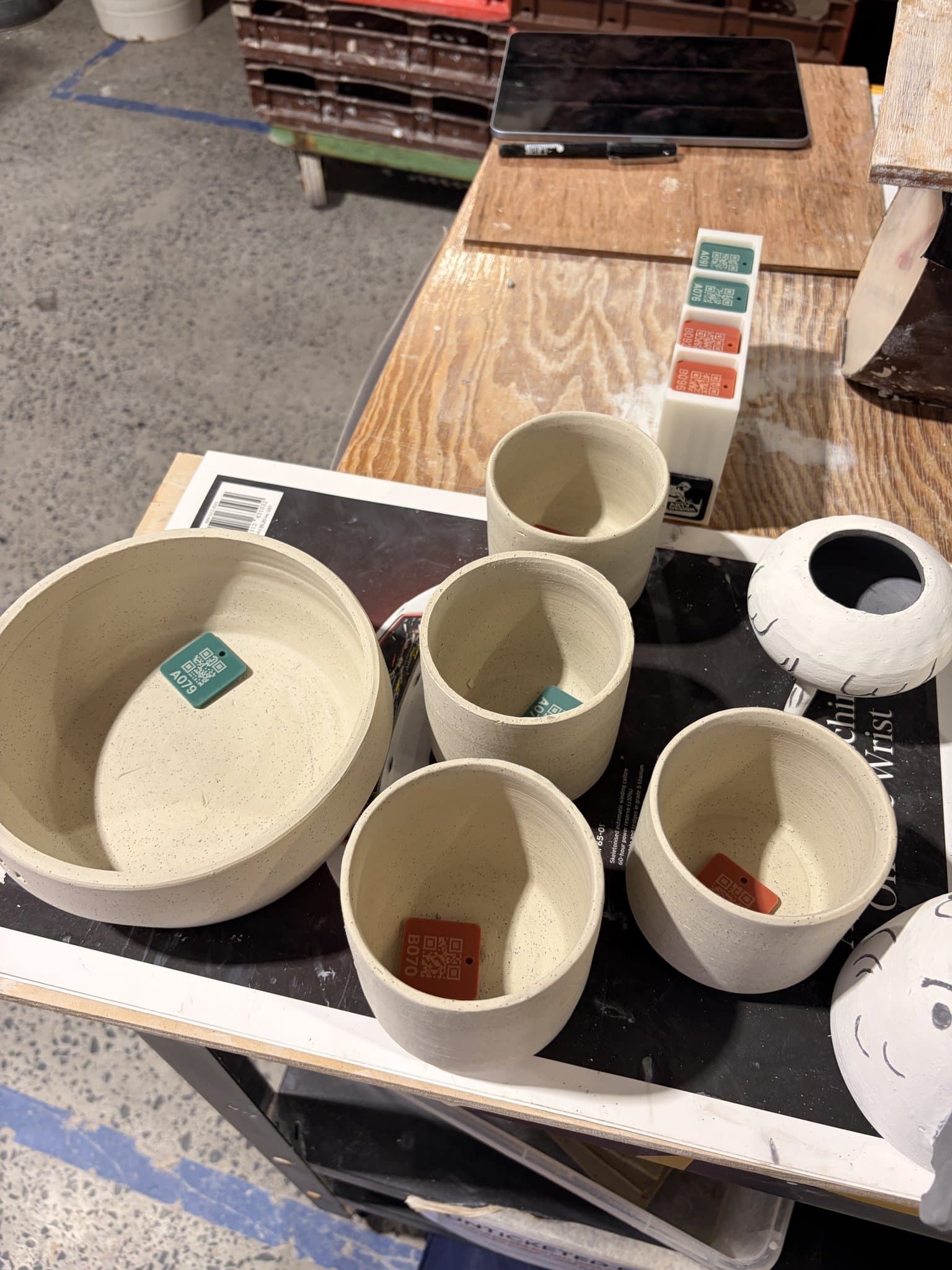 Finished bowls on a studio bench, each tagged with a MudBuddy QR tile.